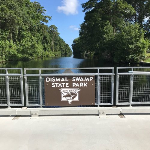 Sign for Dismal Swamp State Park with a view of a calm waterway surrounded by lush greenery.