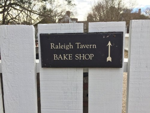Sign on a white fence reading "Raleigh Tavern Bake Shop" with an arrow pointing to the right.