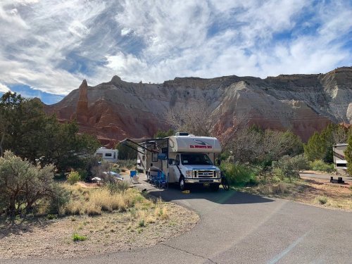 An RV parked at a campsite surrounded by rocky cliffs and greenery under a partly cloudy sky.