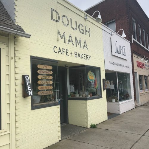 A yellow cafe and bakery named "Dough Mama" with an open sign, next to a shop selling handmade items.