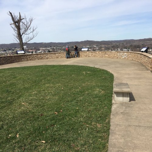 Two people stand on a stone overlook, gazing at a scenic view of a town and hills under a clear sky.