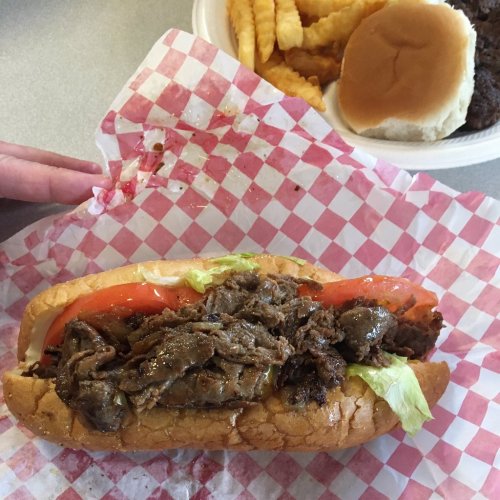 A close-up of a sandwich with sliced beef, lettuce, and tomato on a checkered paper, with crinkle fries and a bun in the background.