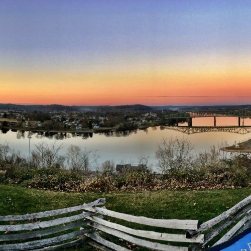 A serene sunset over a river, with a bridge in the distance and trees lining the shore, viewed from a grassy area.
