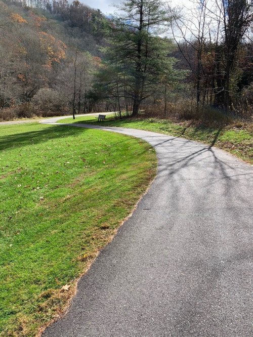 A winding path through a grassy area, with a bench and trees in the background under a partly cloudy sky.