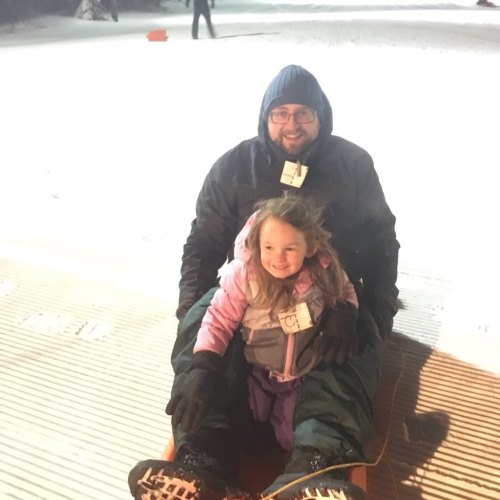 A man and a young girl sit on a sled, smiling, on a snowy slope at night.