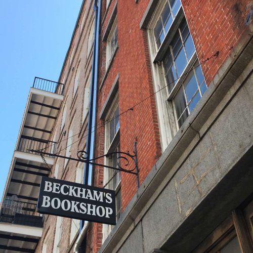 Sign for Beckham's Bookshop hanging from a brick building, with windows visible above and a clear blue sky.
