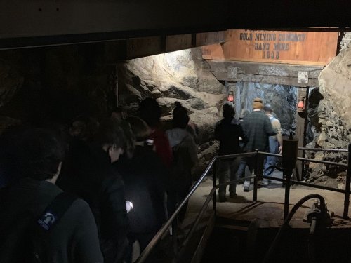 A group of people walking into a dimly lit gold mine, with rock walls and lanterns illuminating the path.