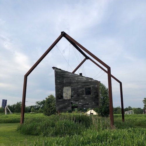 A dilapidated wooden house suspended by a metal frame in a grassy field under a cloudy sky.