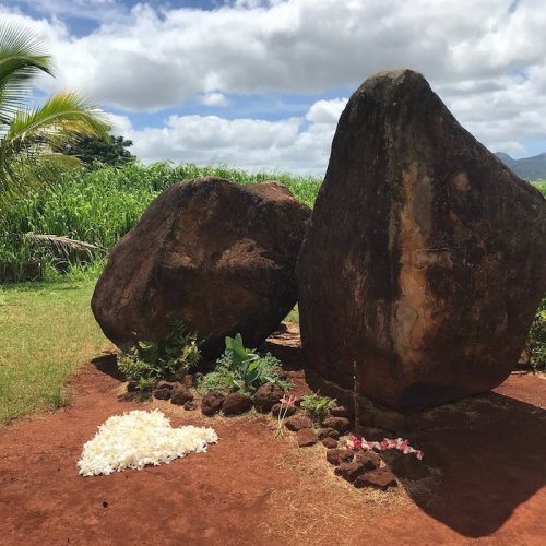 Two large rocks stand in a grassy area, with a heart-shaped arrangement of flowers and stones nearby under a cloudy sky.