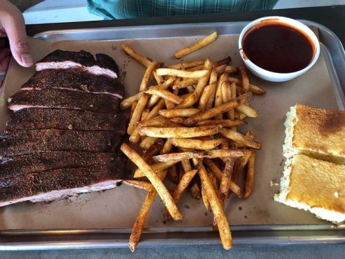 A tray with sliced smoked brisket, seasoned fries, cornbread, and a small cup of barbecue sauce.