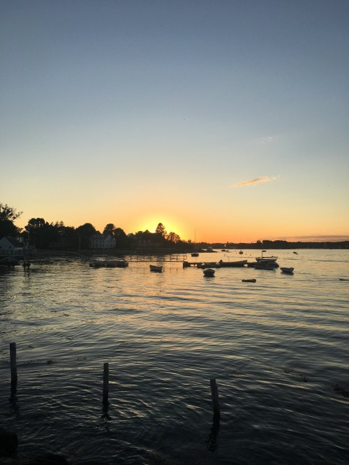 Sunset over a calm waterway, with boats anchored and silhouettes of trees and buildings in the background.