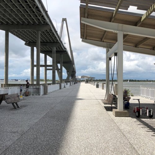 A wide walkway under a bridge, with benches and a view of the water, set against a cloudy sky.