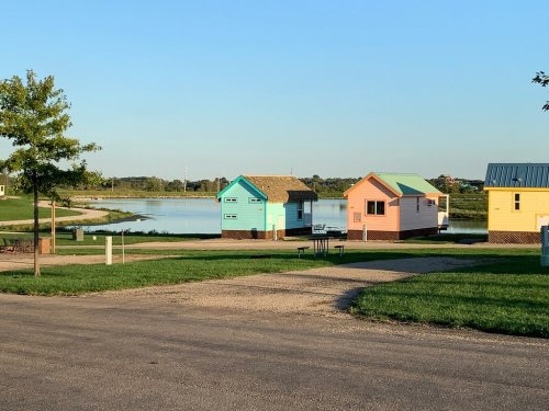 Colorful small houses by a calm lake, surrounded by green grass and trees under a clear blue sky.