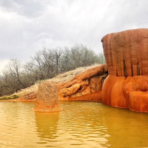 A reddish rock formation rises from a calm, muddy water body, surrounded by sparse trees and a cloudy sky.