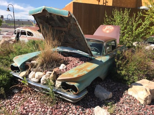 An old, rusted car with a raised hood, overgrown with grass and surrounded by rocks in a neglected area.