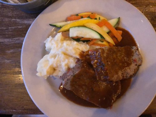 A plate of meatloaf with gravy, mashed potatoes, and a side of mixed vegetables including carrots and zucchini.