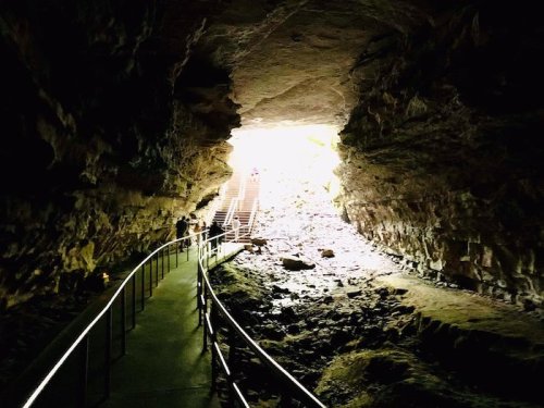 A dimly lit cave entrance with a winding path leading towards bright light and stairs in the background.