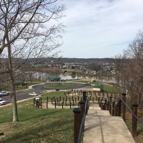 A view from a hillside path overlooking a river and a parking area with trees and buildings in the distance.