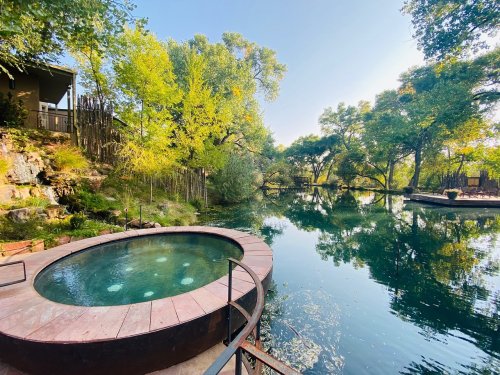 A serene outdoor scene featuring a circular hot tub beside a calm, reflective pond surrounded by lush greenery.