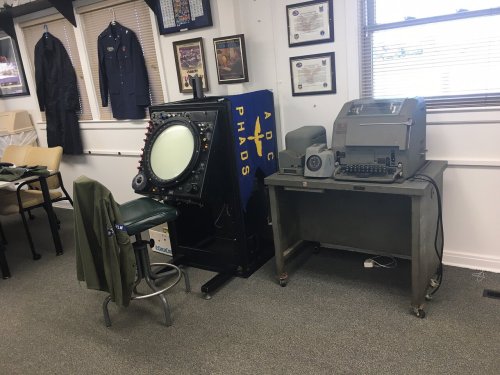 A vintage radar display and an old typewriter sit in a room with military memorabilia and framed certificates.