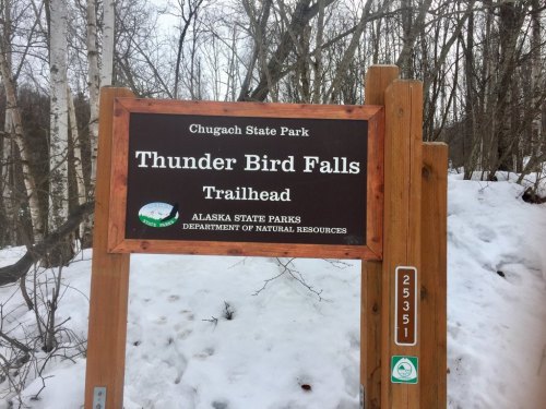 Sign for Thunder Bird Falls Trailhead in Chugach State Park, surrounded by snow and trees.