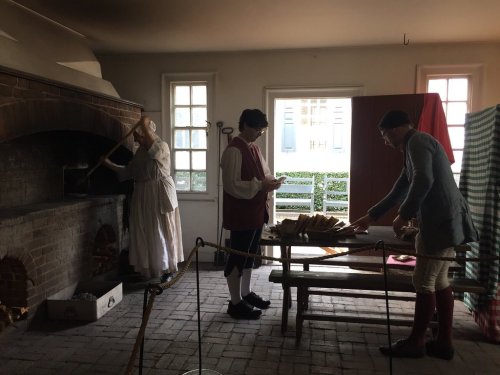 Three people in historical attire work in a rustic kitchen, one tending to a large oven while others prepare food.