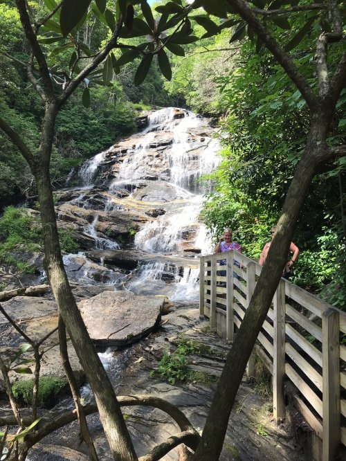 A scenic waterfall cascades down rocky steps, framed by lush greenery and a wooden walkway with visitors nearby.