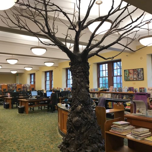 A large, artistic tree sculpture with bare branches stands in a library, surrounded by bookshelves and reading areas.