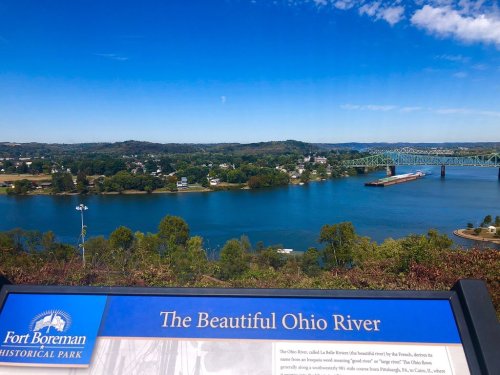 Scenic view of the Ohio River with lush greenery and a bridge in the background, featuring an informational sign.