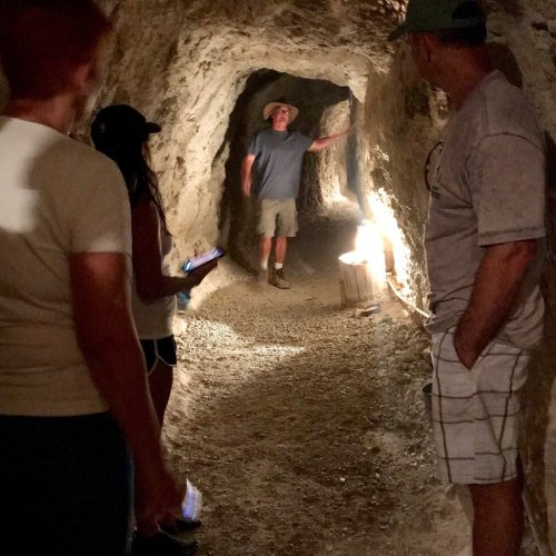 A group of people in a dimly lit cave, with one person standing near a light source, explaining something to the others.