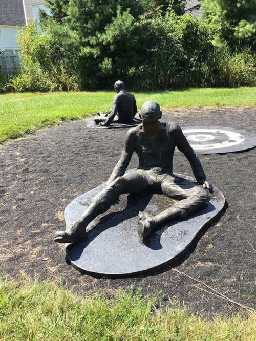 Two bronze sculptures of seated figures on a grassy area, surrounded by dark gravel and greenery.