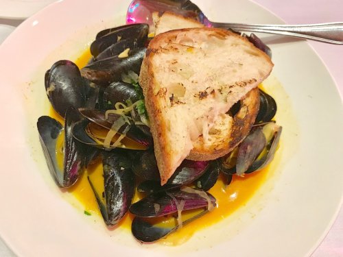 A bowl of mussels in broth, topped with grilled bread and garnished with herbs and onions.