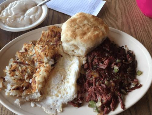 A plate of breakfast featuring hash browns, a fried egg, corned beef hash, and a biscuit, with a side of gravy.