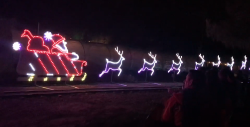 A brightly lit train with a sleigh and reindeer decorations, set against a dark night sky, with spectators watching.