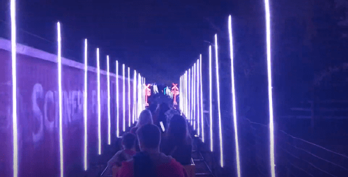 A dimly lit pathway lined with glowing vertical lights, with people walking towards colorful neon signs in the distance.