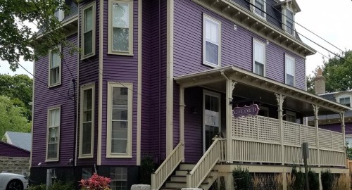 A large purple house with a porch and a sign reading "Gilded," surrounded by greenery and a stone pathway.