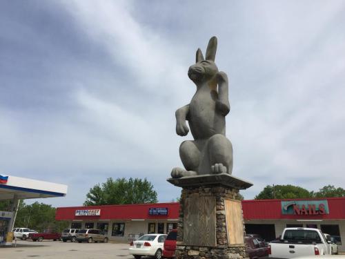 A large stone rabbit statue stands on a pedestal outside a gas station and shops under a cloudy sky.