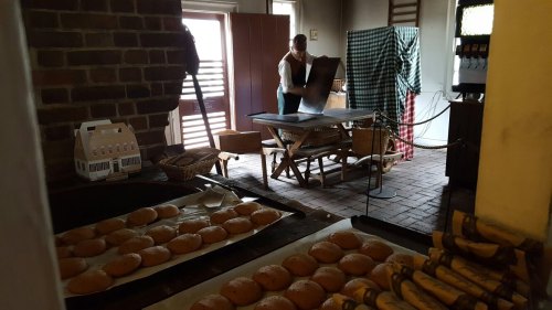A historic kitchen scene with cookies on a tray and a person preparing food at a table in a rustic setting.