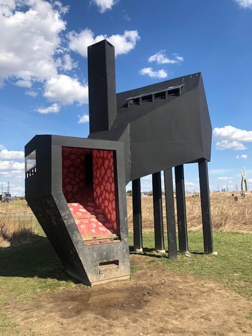 A large, black sculpture resembling a doghouse with red interior, set against a blue sky and grassy landscape.