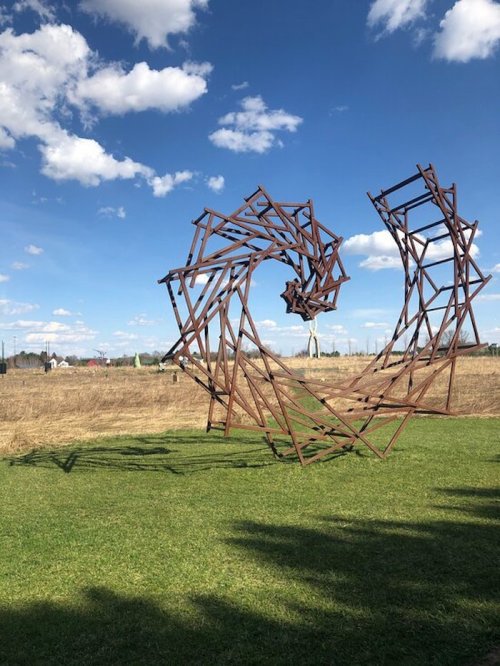 A large, abstract metal sculpture spirals upward against a blue sky, surrounded by grassy fields and distant structures.