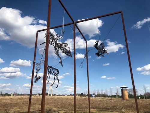 A metal sculpture of suspended animals against a blue sky with clouds, set in an open field.