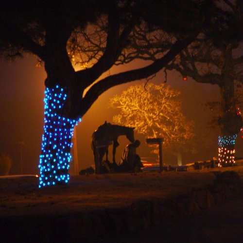 A dimly lit scene featuring a tree wrapped in blue lights, with a silhouette of a person and a horse nearby.