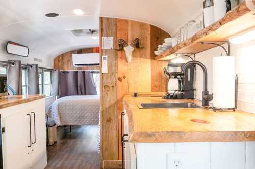 Cozy interior of a renovated bus, featuring a wooden kitchen counter, sink, and a bed area in the background.