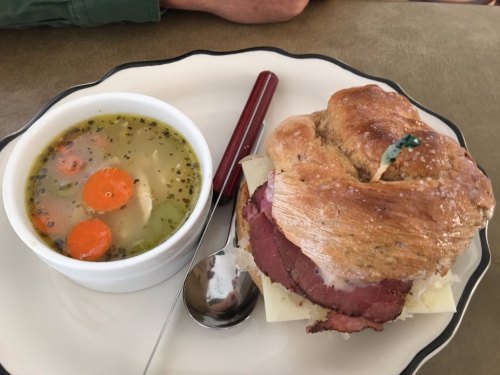A plate with a sandwich on a bun filled with meat and cheese, alongside a bowl of vegetable soup with carrots.