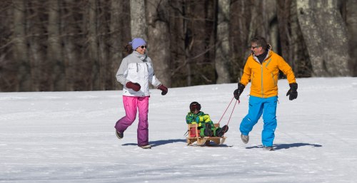 A woman and a man pull a child on a sled through a snowy landscape, surrounded by trees.
