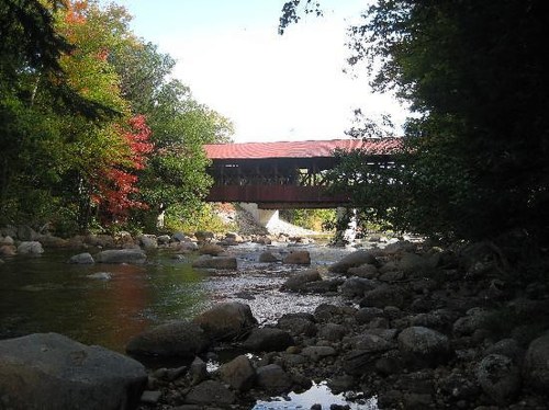 A covered bridge spans a river, surrounded by trees with autumn foliage and rocky riverbanks.