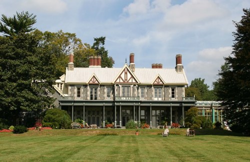 A large, historic house with a gray stone facade, multiple chimneys, and a spacious lawn surrounded by trees.