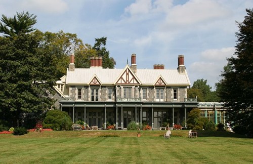 A large, historic house with a gray stone facade and a manicured lawn, surrounded by trees and colorful flower beds.