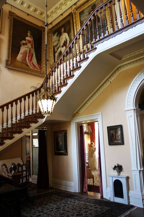 A grand staircase in a historic home, adorned with portraits and elegant decor, leading to a doorway with soft lighting.