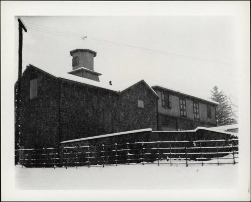 A snow-covered stone building with a tower, surrounded by a fence and trees in a winter landscape.